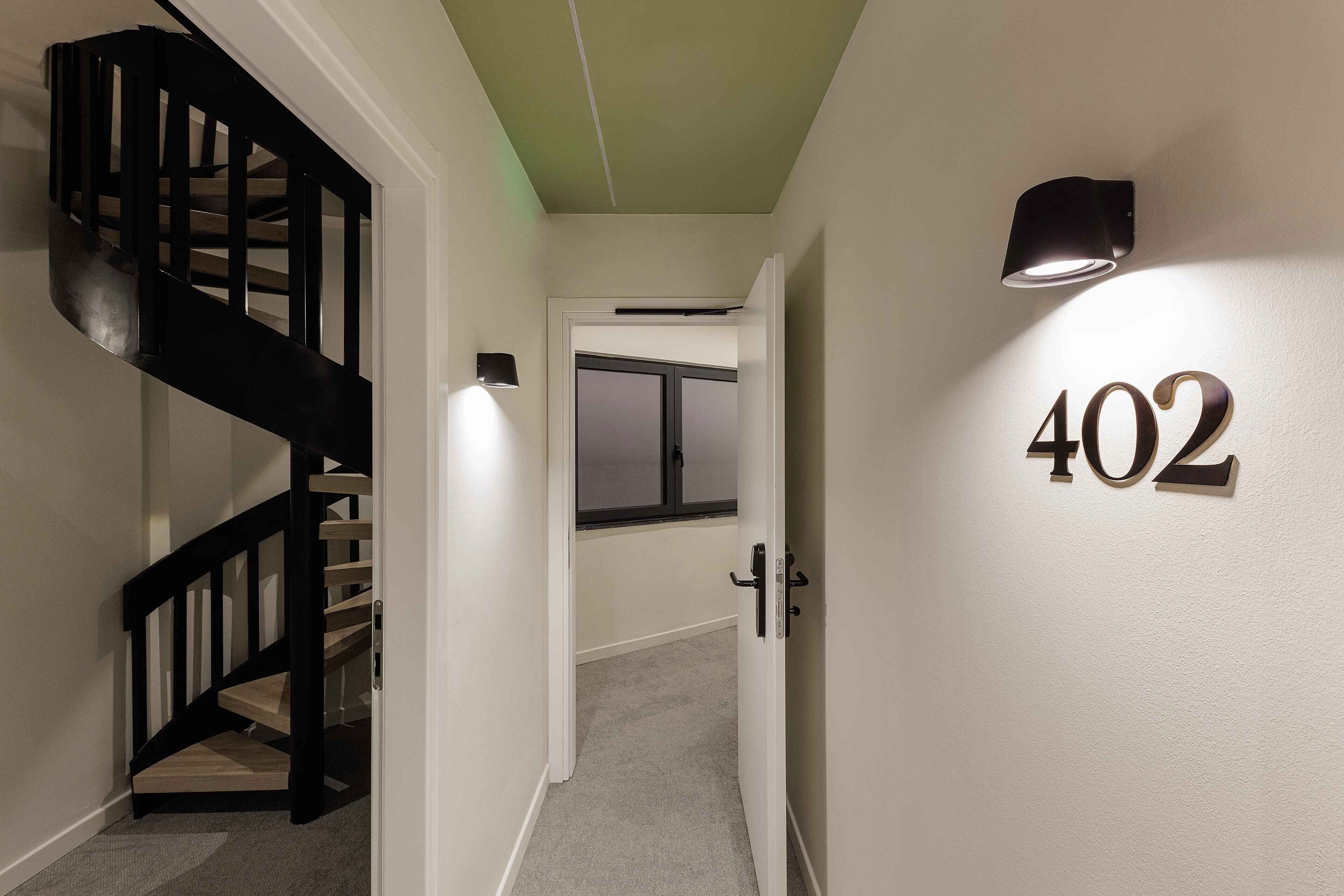 A hallway with a black staircase, a door with the number 402, and a wall-mounted light fixture.