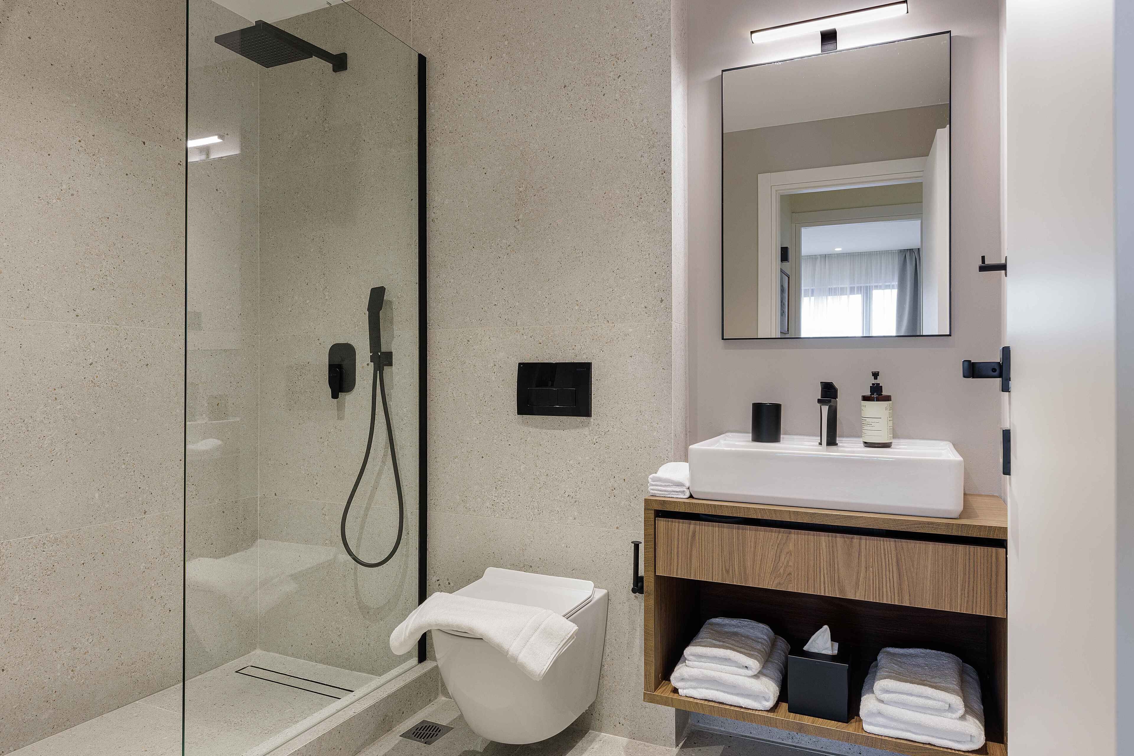 A modern bathroom with a glass-enclosed shower, a white toilet, a wooden vanity with a mirror, and neatly folded towels.