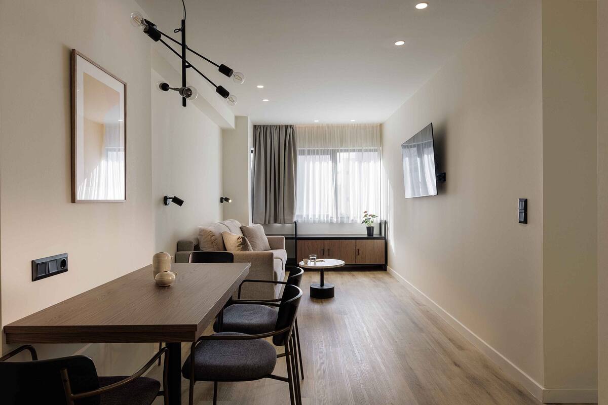 Ein modernes Wohnzimmer mit einem Holzesstisch, dunklen Stühlen, einer Couch und einem an der Wand befestigten Fernseher.