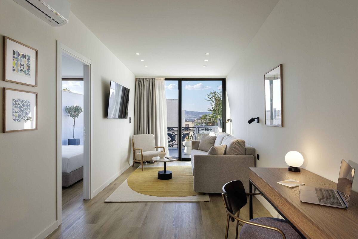 Ein modernes Hotelzimmer mit Holzboden, einer Couch, einem Schreibtisch mit Laptop, einem Fernseher und einem Balkon mit Stadtblick.