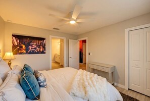 Une chambre à coucher comprenant un lit, des oreillers, une table de chevet, une lampe, un ventilateur de plafond et une porte donnant sur une autre pièce.