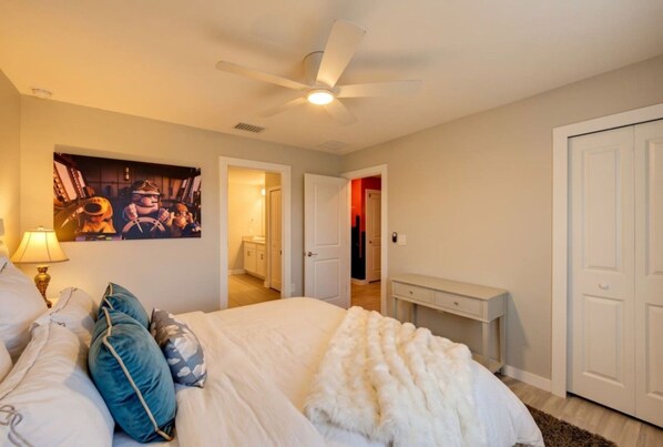 A bedroom with a bed, pillows, a bedside table, a lamp, a ceiling fan, and a door leading to another room.