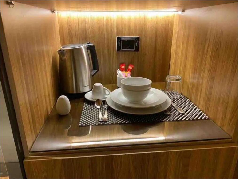 A compact, well-lit room with a wooden finish, featuring a small table set with a teapot, cups, and a bowl, along with a mat and a spoon.