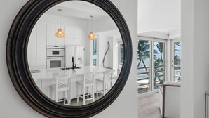 A modern kitchen with white cabinetry and a central island, large windows offering a view of palm trees and the ocean.
