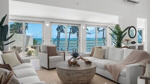 A spacious living room with a large window overlooking a beach and ocean, featuring a white sofa, a round coffee table, and a potted plant.