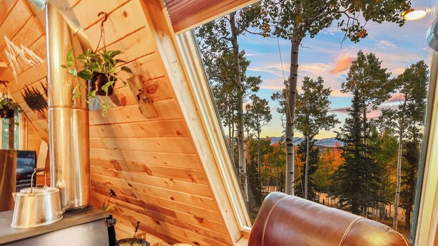 A cozy cabin interior with a wooden interior, a leather couch, a wood stove, and a view of trees and mountains through a large window.