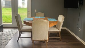 Un comedor con una mesa de madera y cuatro sillas, un televisor montado en la pared y una puerta corrediza de vidrio que da a un jardín trasero.