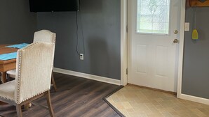 Una habitación con piso de madera oscura, una silla blanca, una mesa de comedor, un televisor de pantalla plana montado en la pared y una puerta con ventanal.