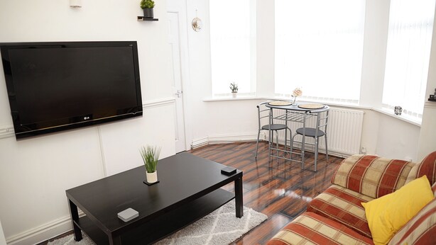 Ein Wohnzimmer mit Flachbildfernseher, einem schwarzen Couchtisch, einem gestreiften Sofa und einer kleinen Essecke mit rundem Tisch und zwei Stühlen.