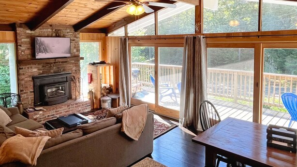 A cozy cabin interior with a fireplace, wooden ceiling beams, a sofa, a dining table, and large windows offering a view of the deck and outdoors. - Pet Friendly Ski House, Game Rm, 4 Mins to Mt Snow (Dover)