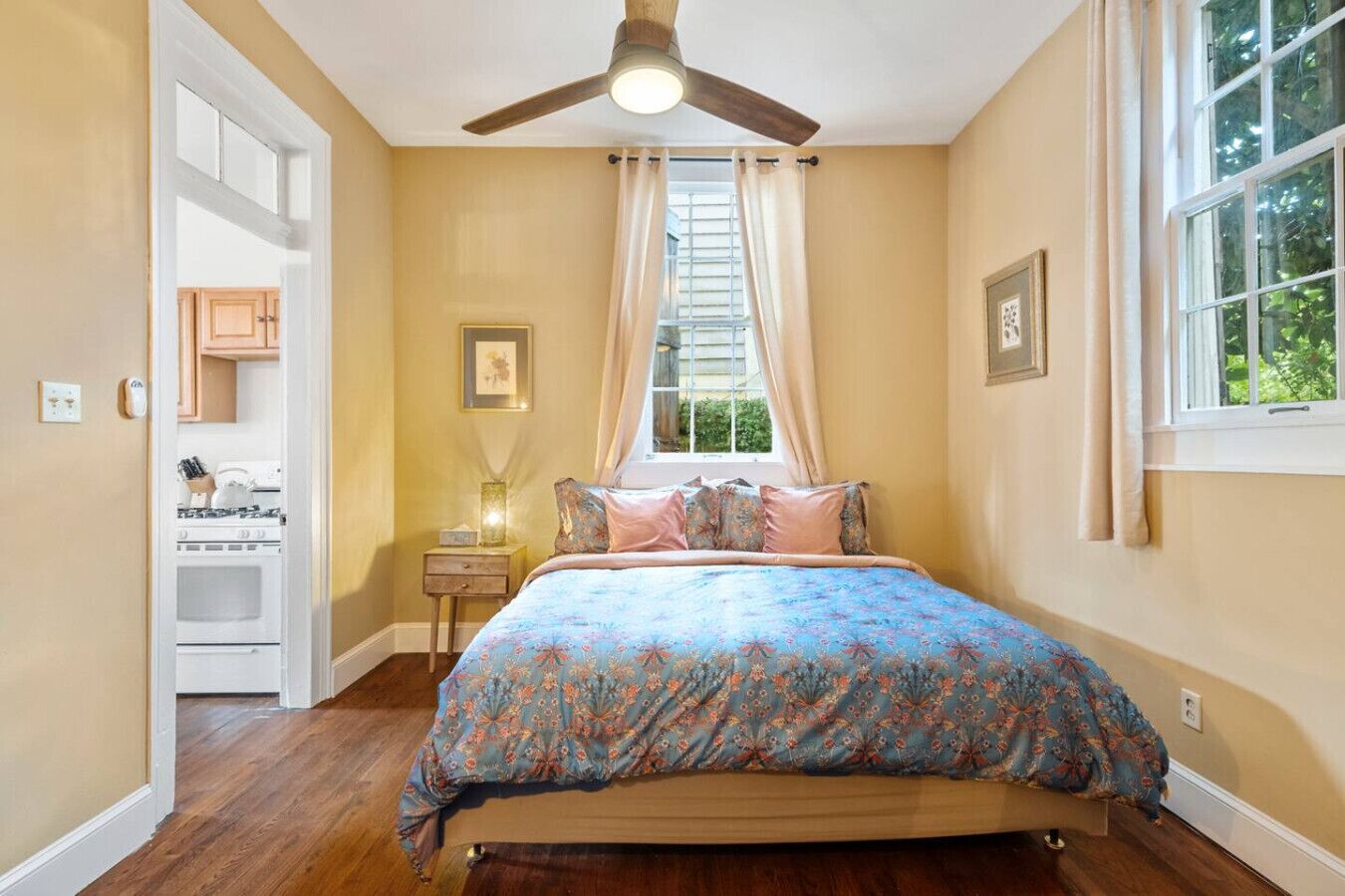 A bedroom with a bed, a ceiling fan, a window with curtains, a nightstand, and a kitchen area visible through an open door.