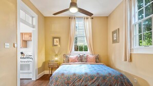 Une chambre à coucher avec un lit, un ventilateur de plafond, une fenêtre avec des rideaux, une table de chevet et un coin cuisine visible à travers une porte ouverte.