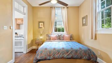 A bedroom with a bed, a ceiling fan, a window with curtains, a nightstand, and a kitchen area visible through an open door.