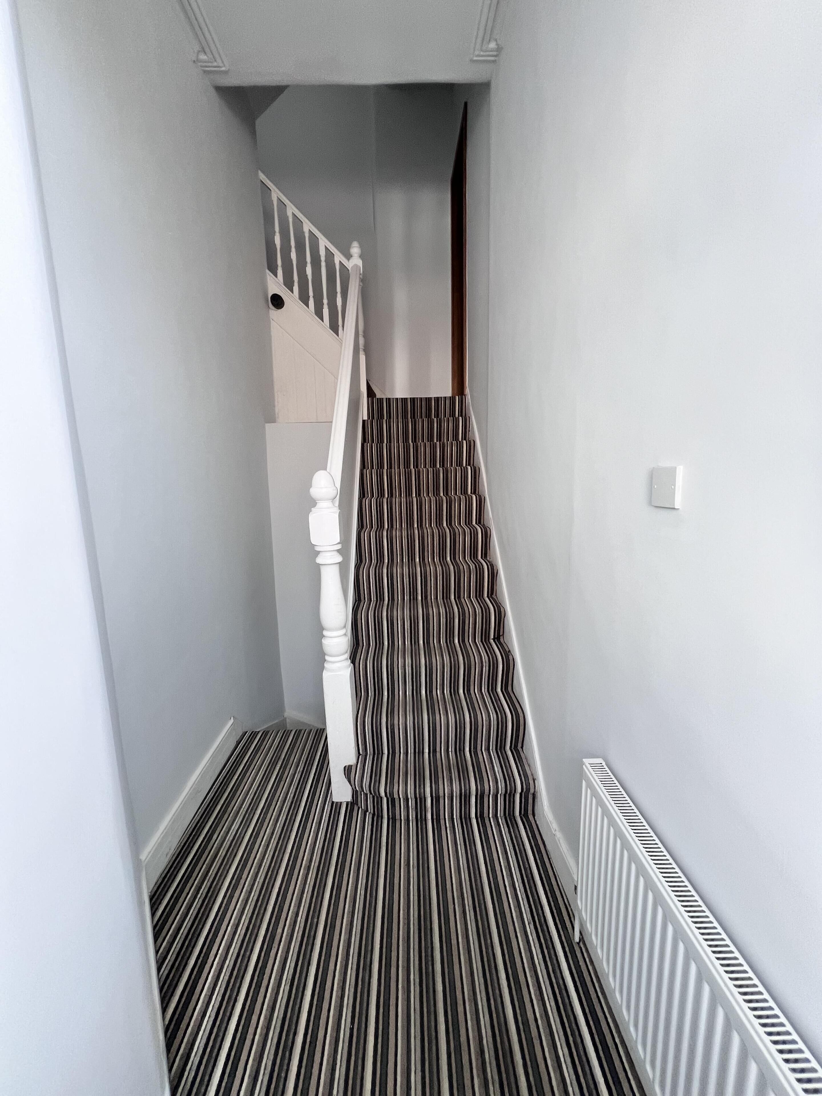 A narrow hallway with a striped carpet, a white staircase with a wooden handrail, and a white radiator on the wall.