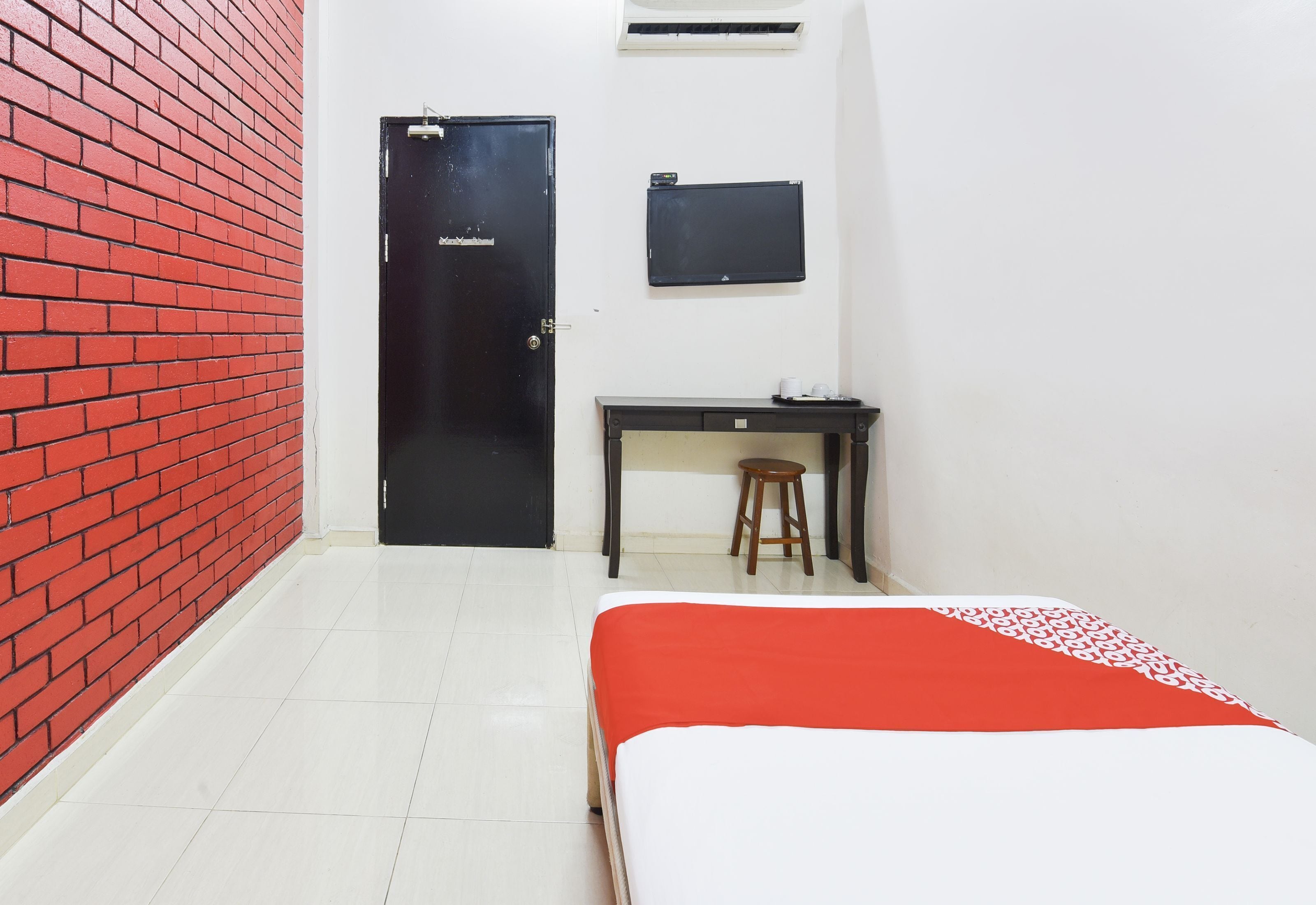 A room with a red brick wall, a black door, a desk with a stool, and a bed with a red and white bedspread.