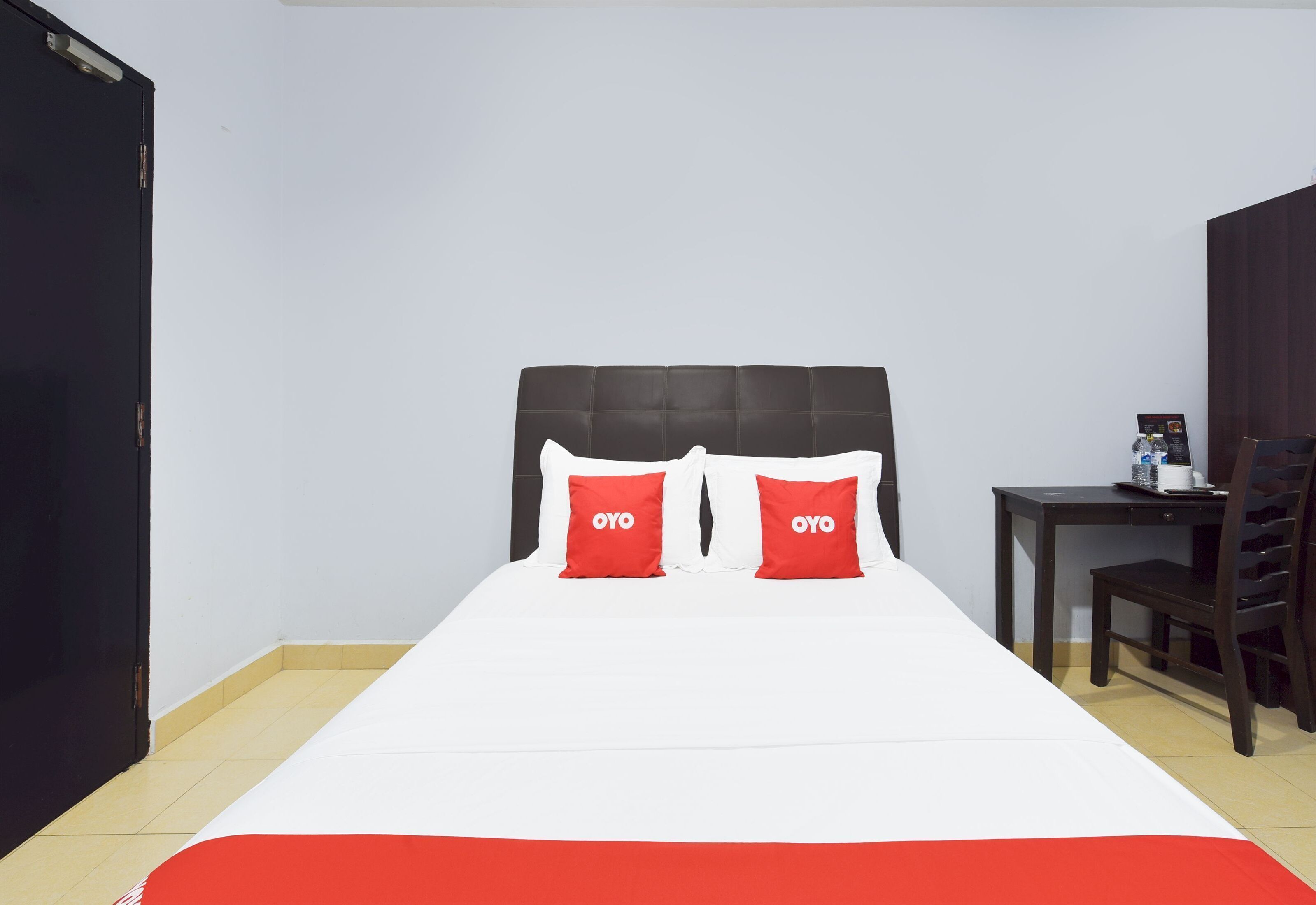 A hotel room with a bed, two red pillows, a wooden headboard, a small table with a chair, and a black door.