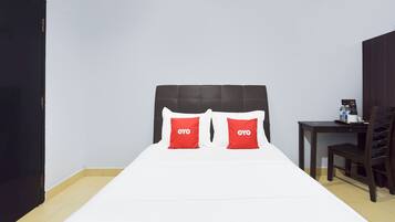 A hotel room with a bed, two red pillows, a wooden headboard, a small table with a chair, and a black door.