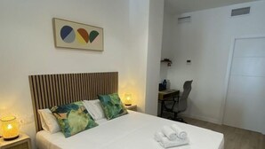 Una habitación de hotel moderna con una cama, una pequeña zona de estar con cojines, un estante en la pared y un elemento decorativo en la pared.