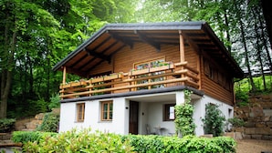 Een houten hut met een balkon, omgeven door weelderige begroeiing en stenen muren.
