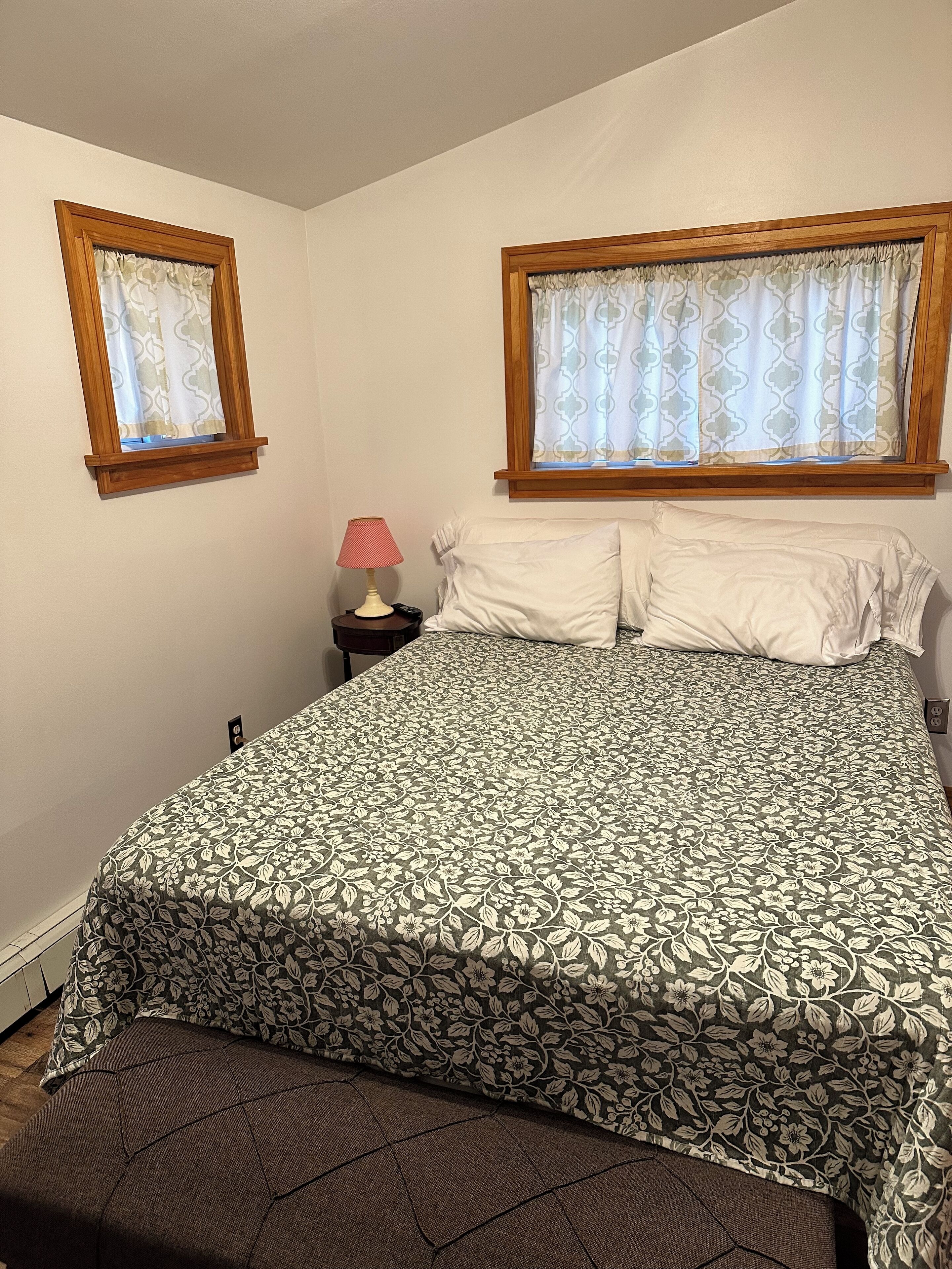 Un dormitorio con una cama, dos ventanas con cortinas, una mesita de noche con una lámpara y un reposapiés marrón.