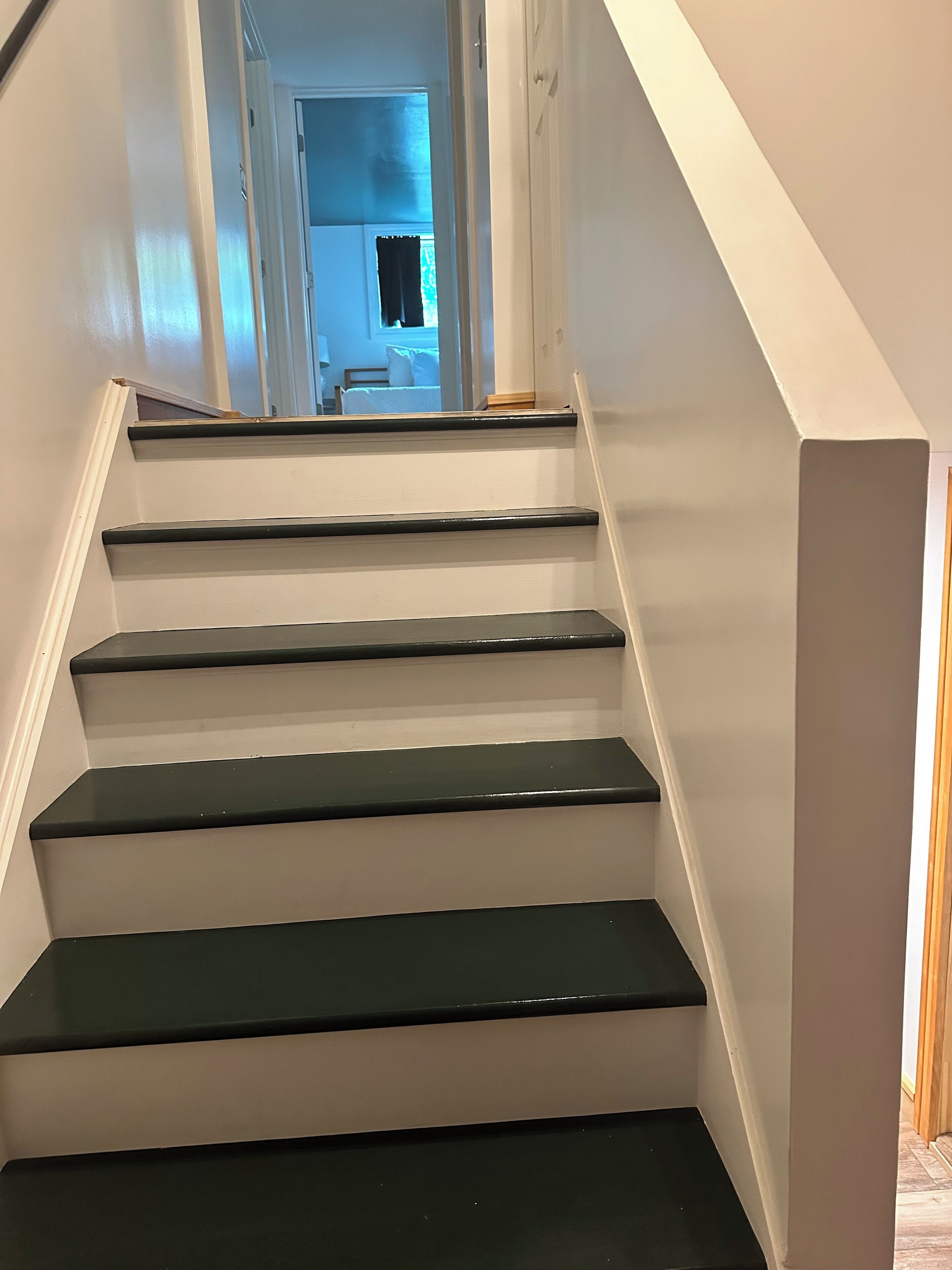 A staircase with white walls and dark steps leading to a room with a bed and a window.