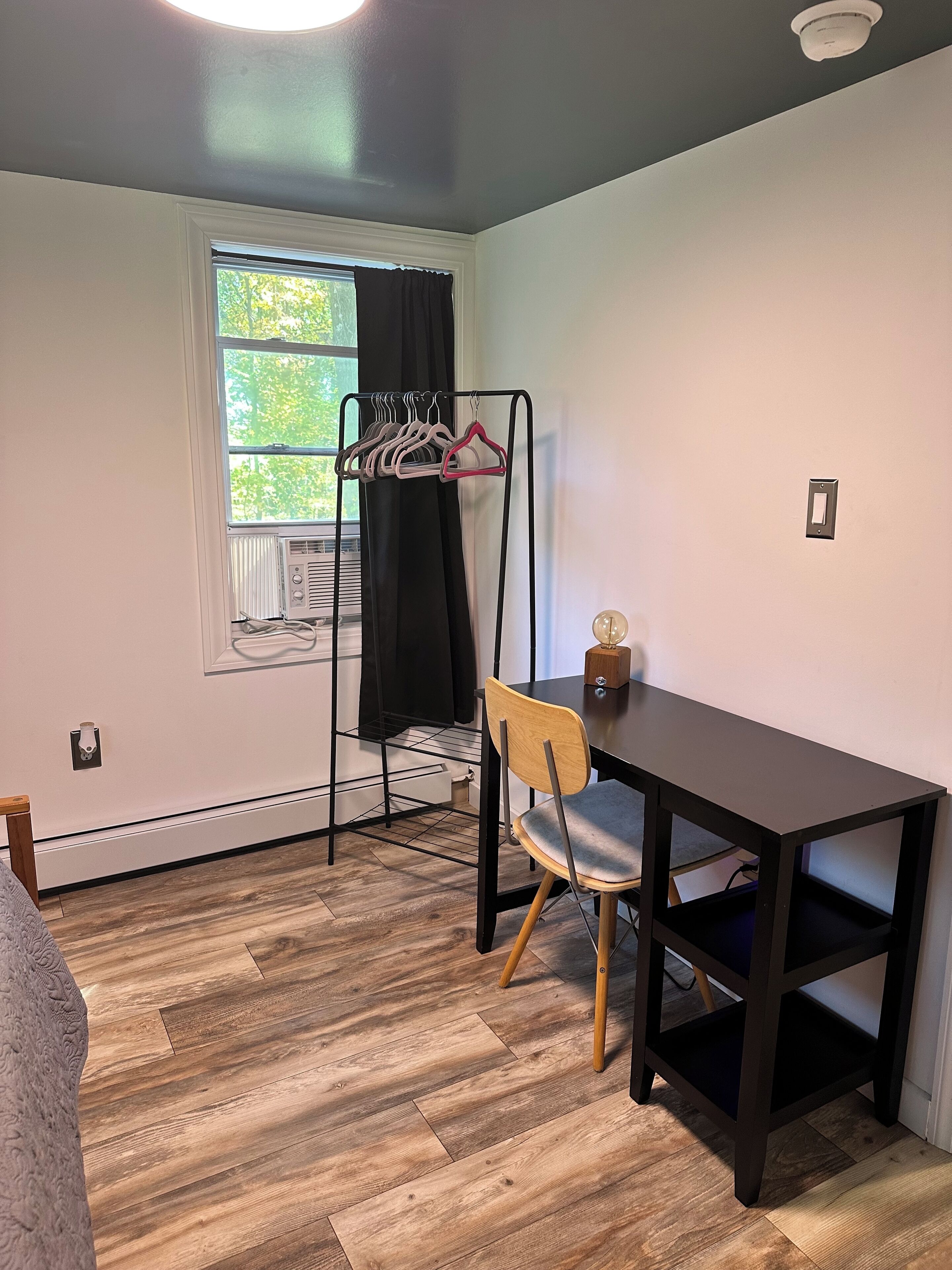 Una habitación con piso de madera, un escritorio negro, una silla, perchero con colgadores, ventana con cortinas y una lámpara en el techo.