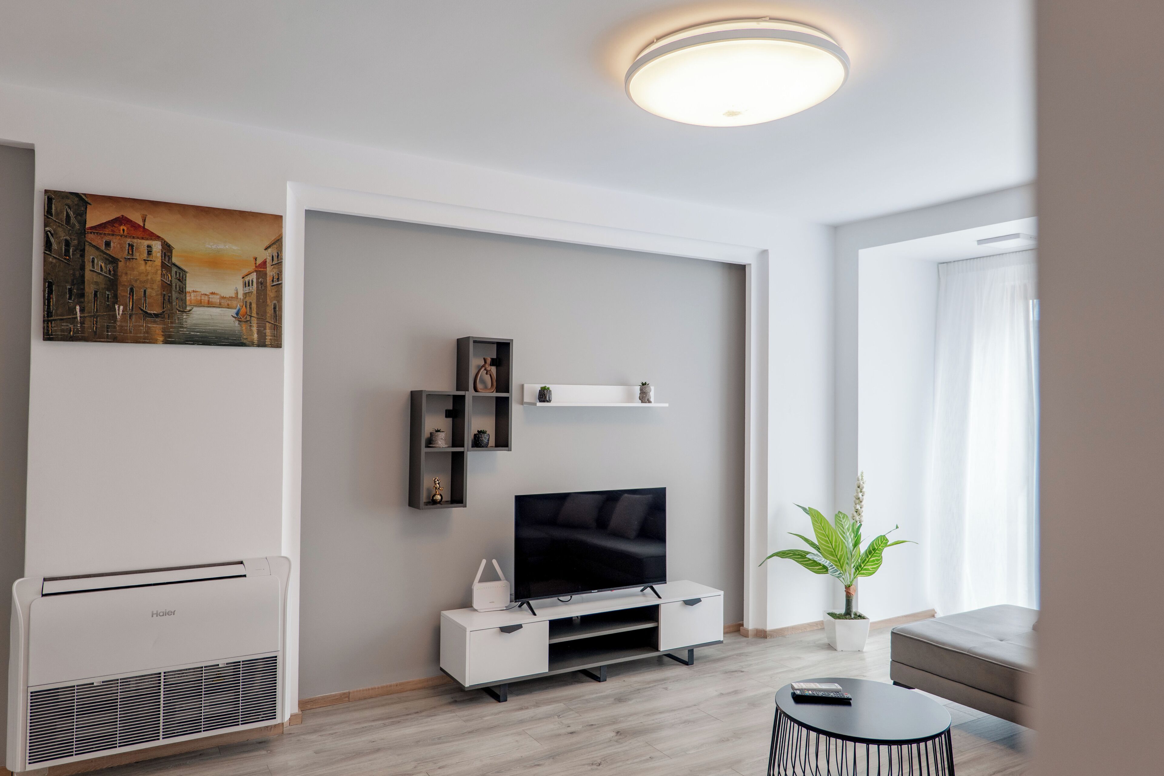 A modern living room with a flat-screen TV, white furniture, a mounted picture, and a ceiling light fixture.
