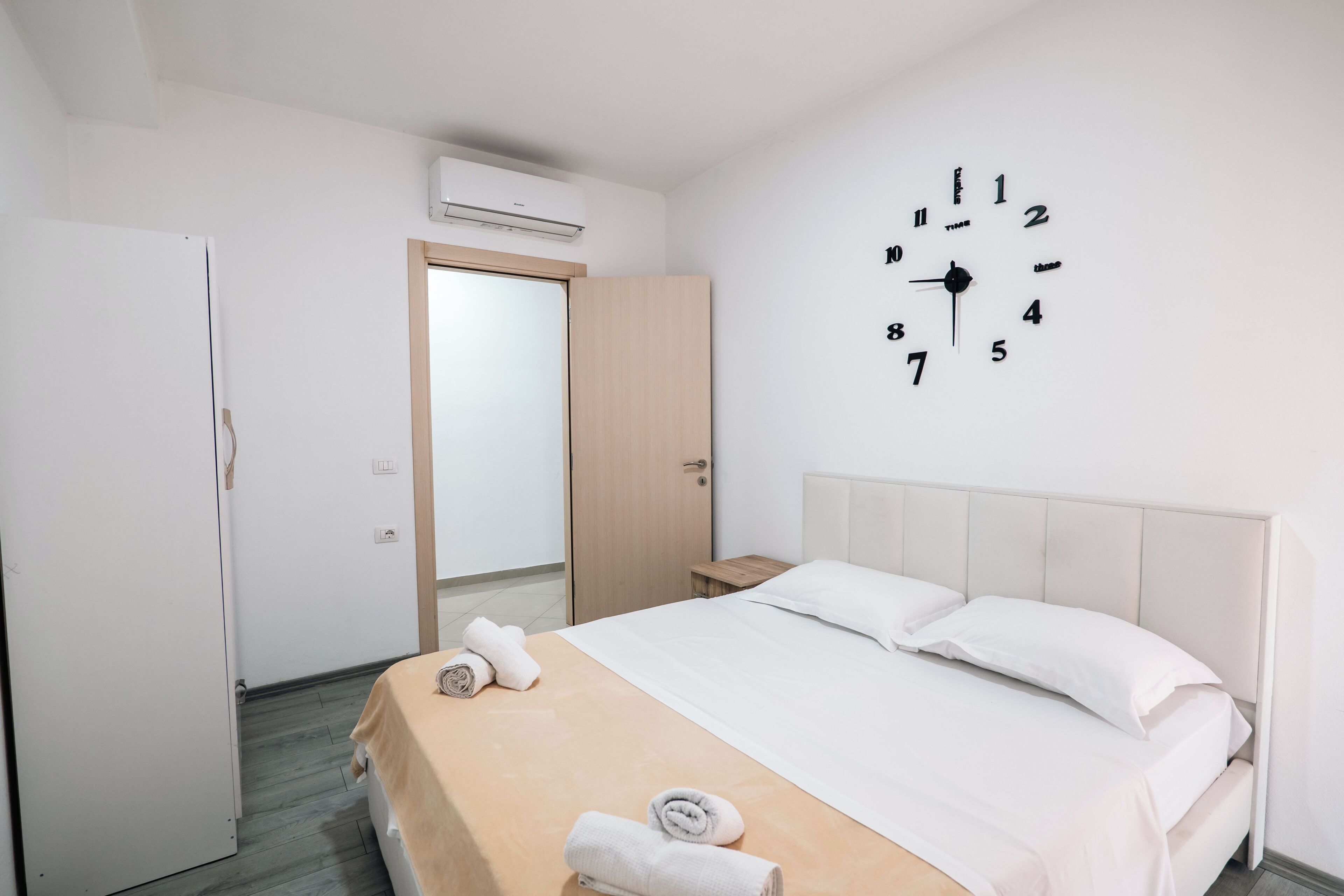 A hotel room with a bed, a wall clock, a door, and an air conditioning unit.