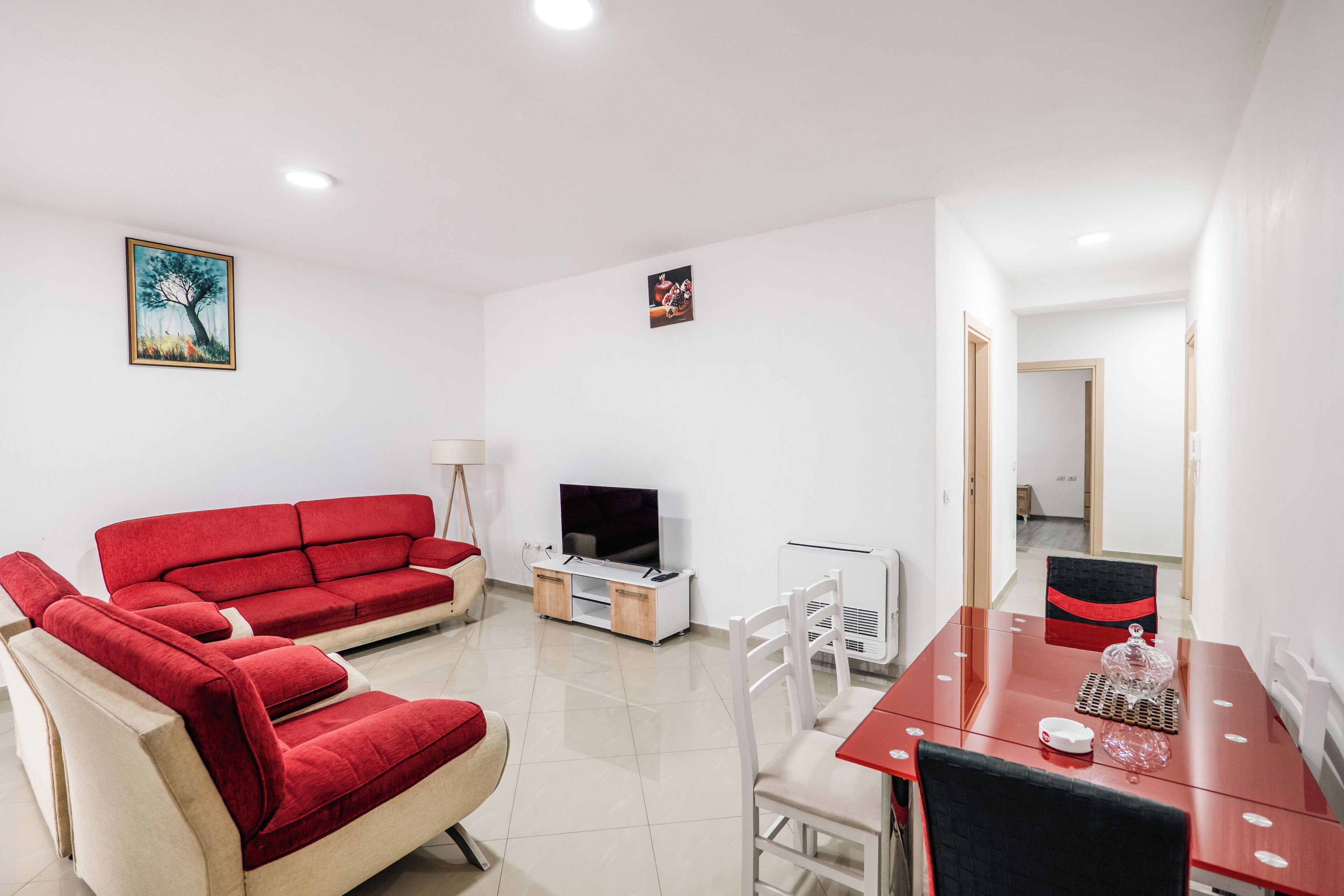Un salon avec un canapé rouge, un téléviseur, une table à manger avec des chaises et un couloir menant à une autre pièce.