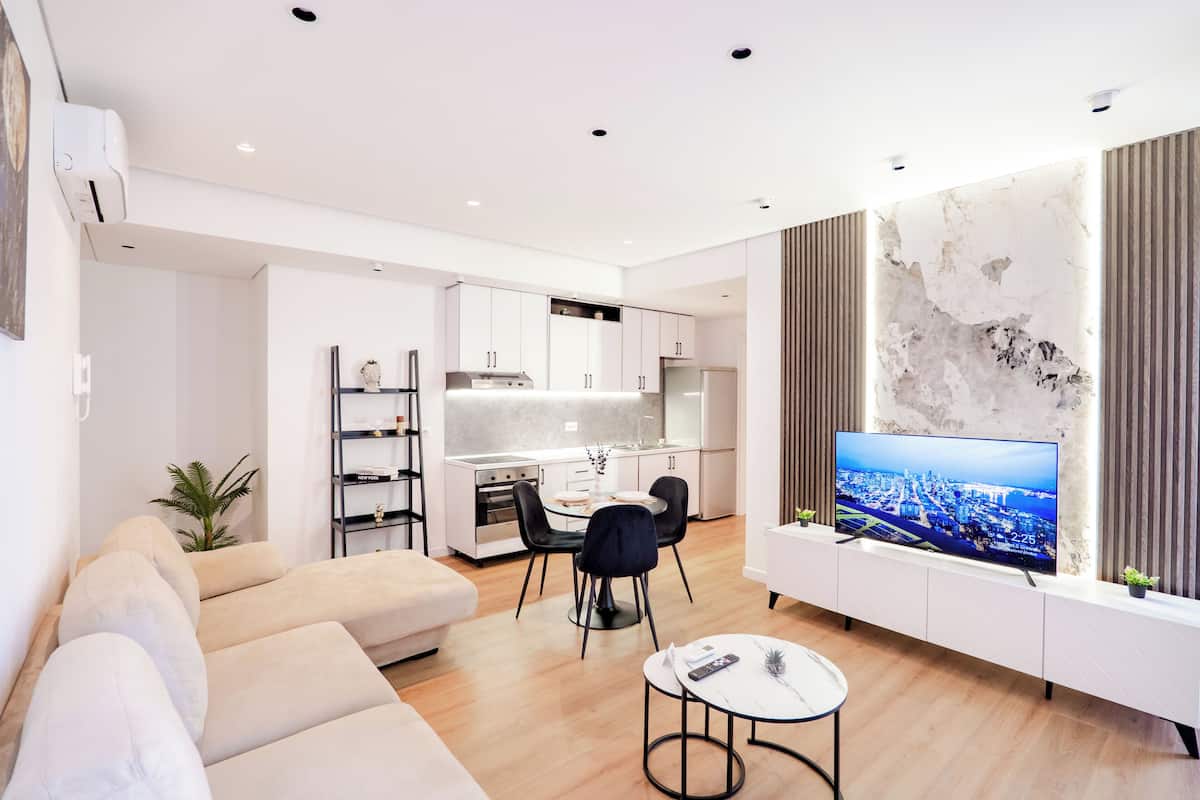 Un salon moderne avec un canapé, une table à manger et des chaises, un téléviseur fixé au mur et une cuisine en arrière-plan.