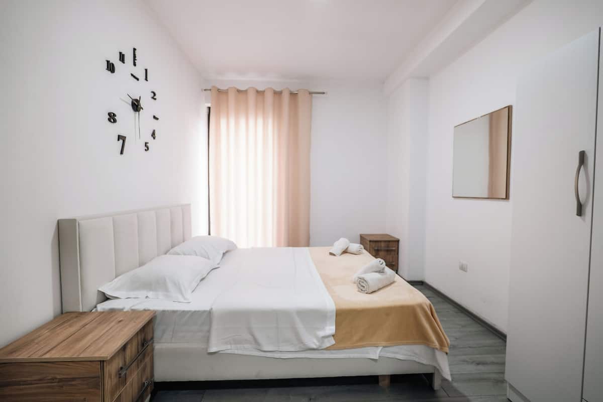 Une chambre à coucher avec un lit, une table de chevet en bois, une horloge murale, un miroir et une fenêtre avec des rideaux.