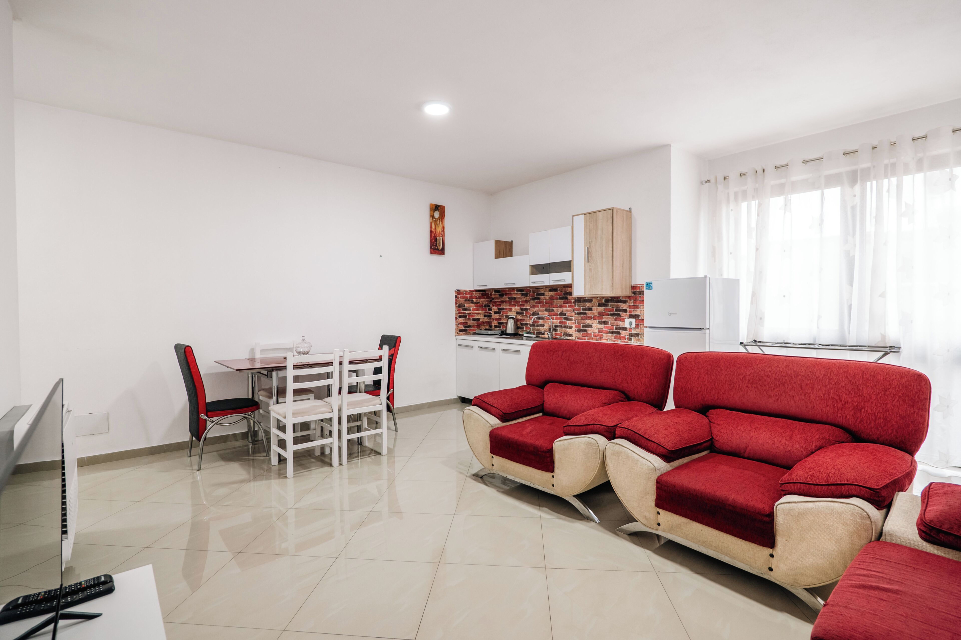 Una sala de estar moderna con un sofá rojo, un comedor con una mesa y sillas blancas, y una cocina con gabinetes blancos y salpicadero de ladrillo.