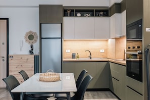 Une cuisine compacte équipée d’un réfrigérateur, d’un four à micro-ondes et d’un four traditionnel, d’une table à manger avec des chaises et d’un petit meuble sur lequel est apposée une étiquette indiquant « DOMICILE ».