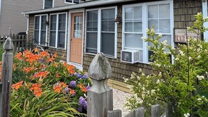 Et tosyltet hus med en overdækket veranda, et træhegn og mange blomstrende planter.