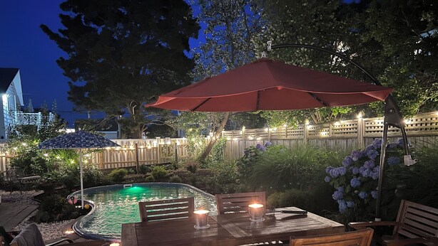 En udendørs patio med pool, træmøbler og en rød parasol.
