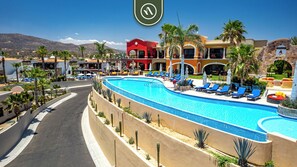 A resort with a large swimming pool, sun loungers, palm trees, and a mountain backdrop. - Hacienda Encantada by Naya Homes (Cabo San Lucas)