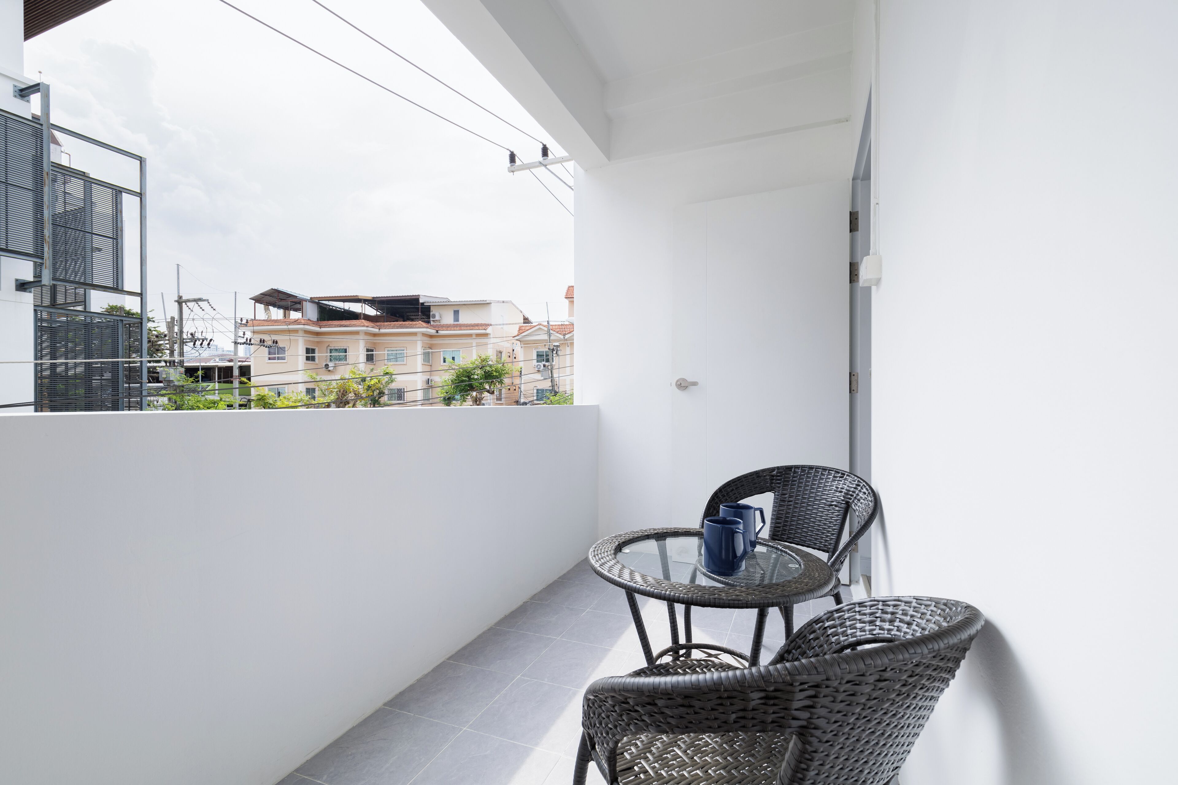 A balcony with a glass table and wicker chairs, overlooking a cityscape with buildings and greenery.