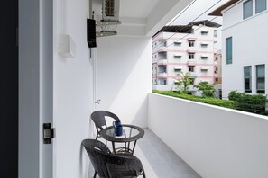 Een balkon met een kleine tafel en twee stoelen, een wandairconditioner en uitzicht op een meerlaags gebouw met balkons.