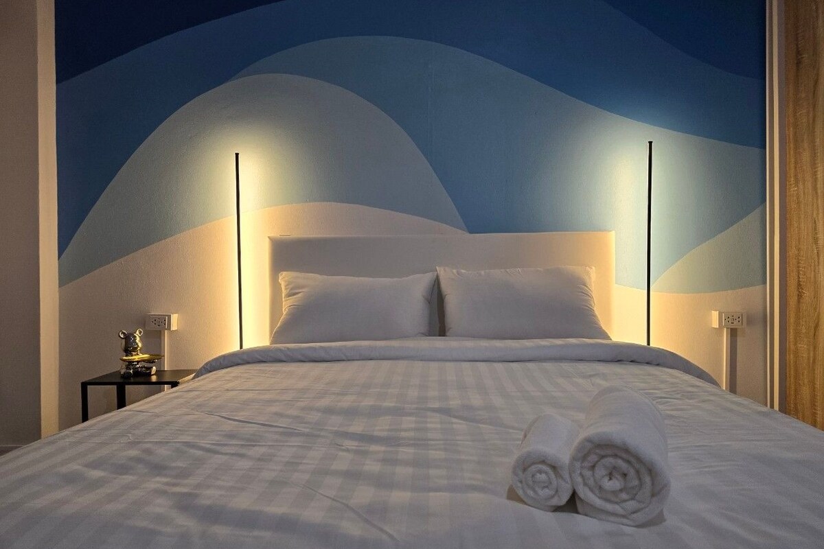 Una cama bien hecha con sábanas blancas y toallas enrolladas, una mesita de noche con lámpara, y una pared con diseño ondulado.
