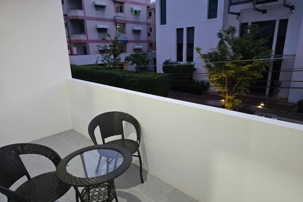 A balcony with two wicker chairs and a glass table, overlooking a residential area with buildings and greenery.