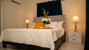 Una cama bien tendida con colcha blanca, cabecera gris con botoneras y un florero con flores amarillas. Hay dos mesitas de noche con lámparas, y una mesita de noche oscura con cajones.