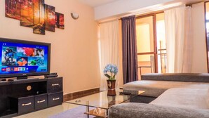 A hotel room with a flat-screen TV, a glass coffee table, a sofa, a bed, a vase with flowers, and a window with curtains. - Lux Suites Makao Rossyln Apartments (Nairobi)