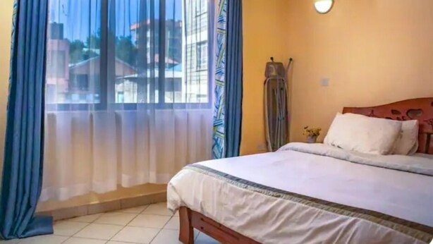A bedroom with a bed, curtains, a wall-mounted light, and a window with a view of buildings. - Lux Suites Makao Rossyln Apartments (Nairobi)