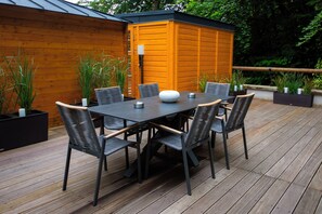 Une terrasse en bois avec une table et des chaises, entourée de plantes en pot et d’une structure en bois.