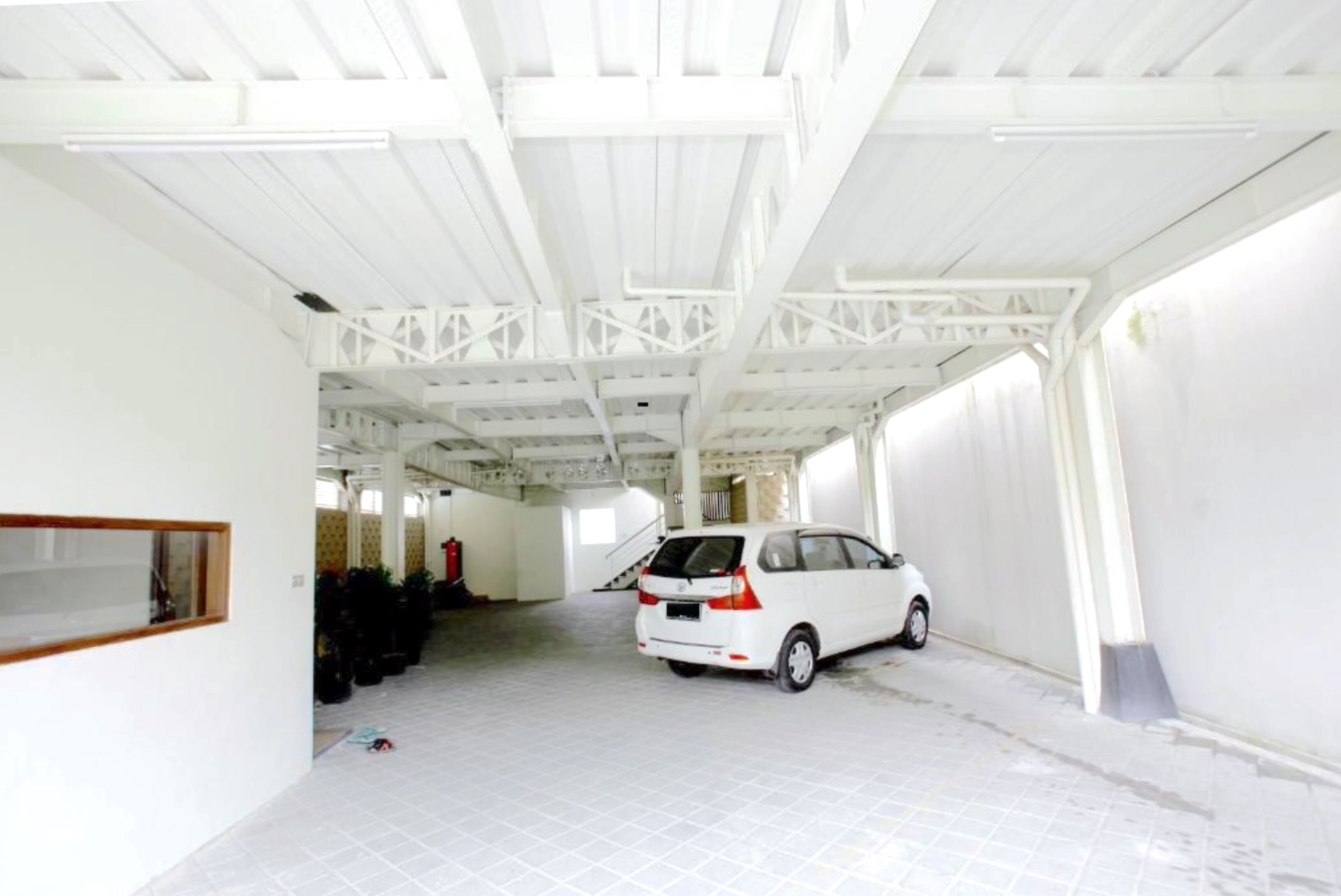 Um amplo espaço interno com um carro branco estacionado no centro, uma escada que leva a um andar superior e uma grande janela à esquerda.