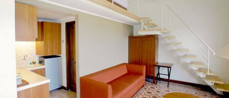 A modern living room with a sofa, a small table, a kitchen area with wooden cabinets, and a staircase.