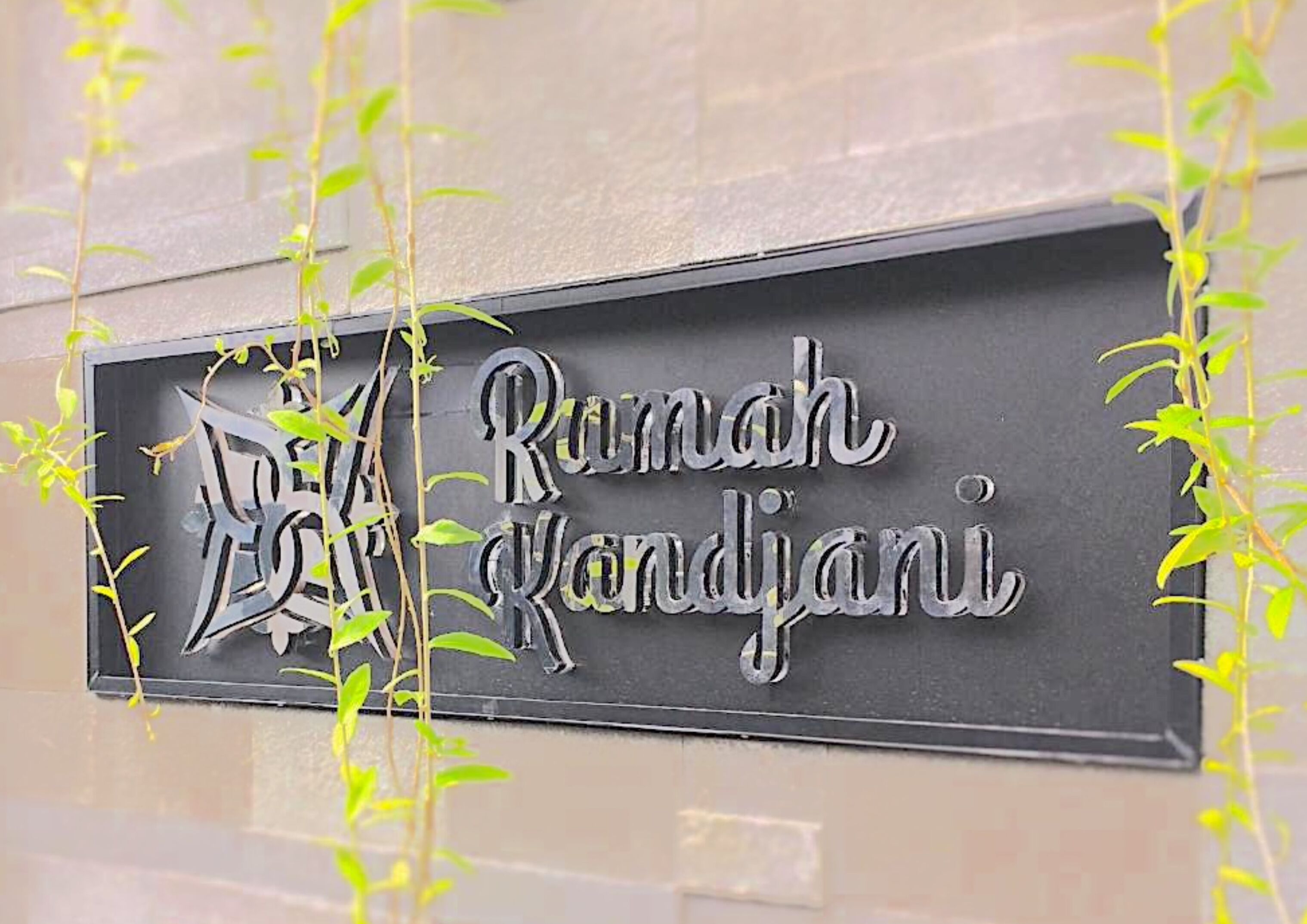 A sign with the text "Rumah Kandjari" on a wall, surrounded by green plants.