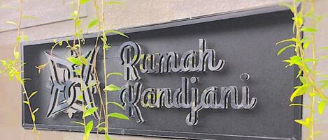 A sign with the text "Rumah Kandjari" on a wall, surrounded by green plants.