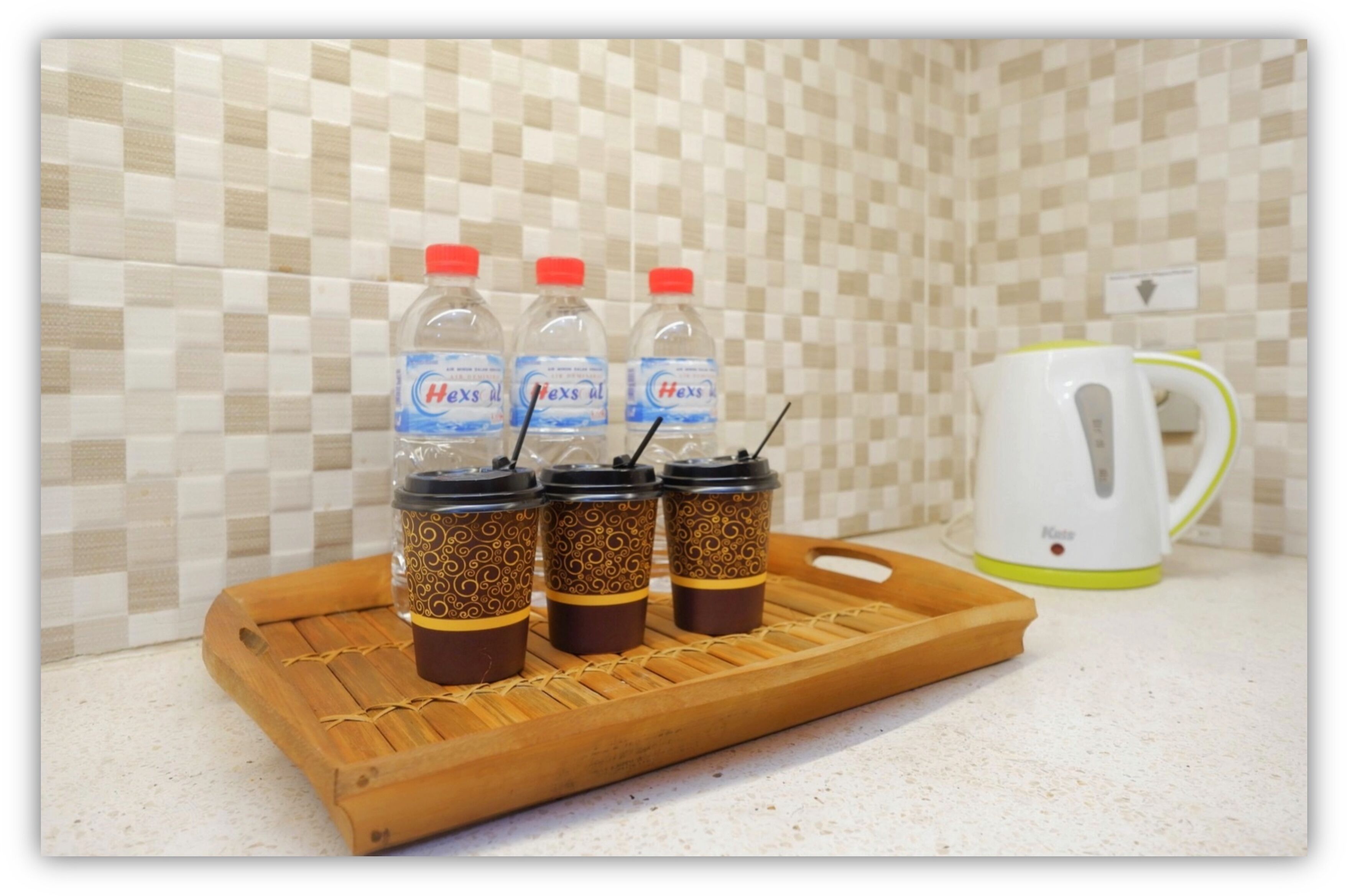Un comptoir de cuisine sur lequel se trouve un plateau contenant quatre bouteilles d’eau et trois tasses de café, une bouilloire blanche et un pare-éclaboussures carrelé.
