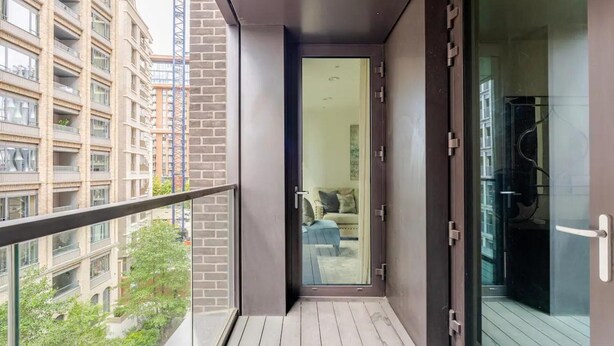 A balcony with a view of a cityscape, a glass door leading to a room with a bed and a chair, and a wooden floor. - Luxury Riverfront Retreat in London (London)